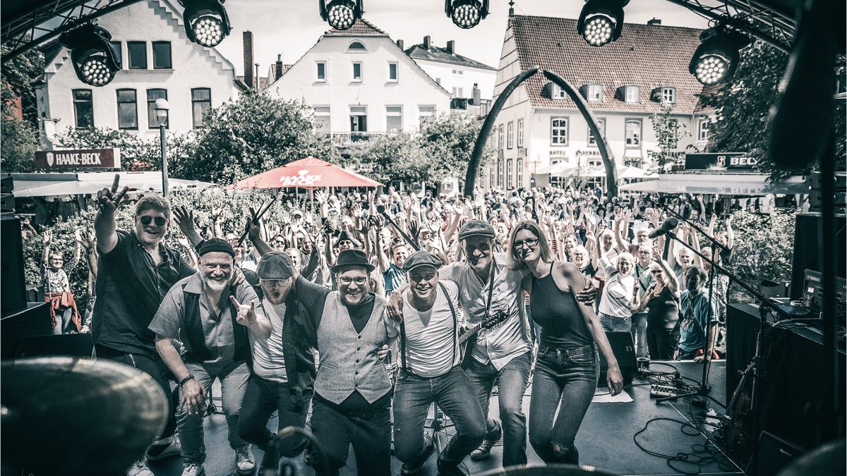 Eine Musikgruppe steht auf einer Bühne. Vor der Bühne stehen ganz viele Menschen