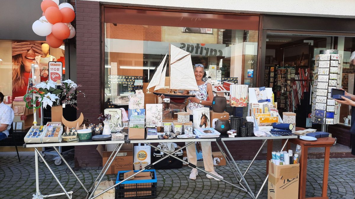 Eine Frau steht hinter einem Flohmarkt Tisch und hält ein kleines Schiff hoch